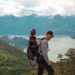 A father carries his child on a scenic mountain trail, embracing adventure and parenthood.